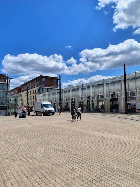 A wide view of the exterior of the Bentall Centre in Kingston upon Thames under a bright blue sky with scattered white clouds. In the foreground, there is a large paved pedestrian area with several people walking, some pushing strollers or carrying bags. A white delivery van is parked near the building's entrance, alongside traffic cones and a few pedestrians nearby. The building has a modern glass and steel façade, with large windows and a transparent canopy over the entrance. Adjacent to the main structure are residential or office buildings with brick and glass exteriors. This scene illustrates an urban setting suitable for home relocation activities, with visible space for loading and unloading furniture or boxes during moving or packing processes, supported by the presence of a moving company like manwithavankingstonuponthames.co.uk.