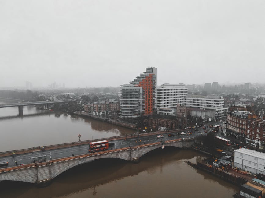 Aerial view of Kingston upon Thames on a cloudy day, showing a prominent modern residential building with white and orange exterior, situated near the River Thames. A bridge crosses the river, with vehicles including a red and white bus and cars traveling across it. Adjacent to the riverbank are various low-rise brick and concrete buildings, with some streets visible around the area. The scene depicts urban traffic and typical cityscape elements, consistent with a location involved in house removals and furniture transport services as part of home relocation routines carried out by companies such as manwithavankingstonuponthames.co.uk.