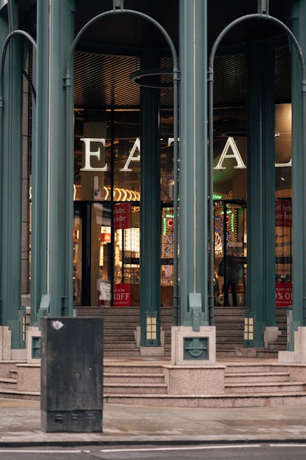 A wide view of the exterior of the Bentall Centre in Kingston upon Thames under a bright blue sky with scattered white clouds. In the foreground, there is a large paved pedestrian area with several people walking, some pushing strollers or carrying bags. A white delivery van is parked near the building's entrance, alongside traffic cones and a few pedestrians nearby. The building has a modern glass and steel façade, with large windows and a transparent canopy over the entrance. Adjacent to the main structure are residential or office buildings with brick and glass exteriors. This scene illustrates an urban setting suitable for home relocation activities, with visible space for loading and unloading furniture or boxes during moving or packing processes, supported by the presence of a moving company like manwithavankingstonuponthames.co.uk.
